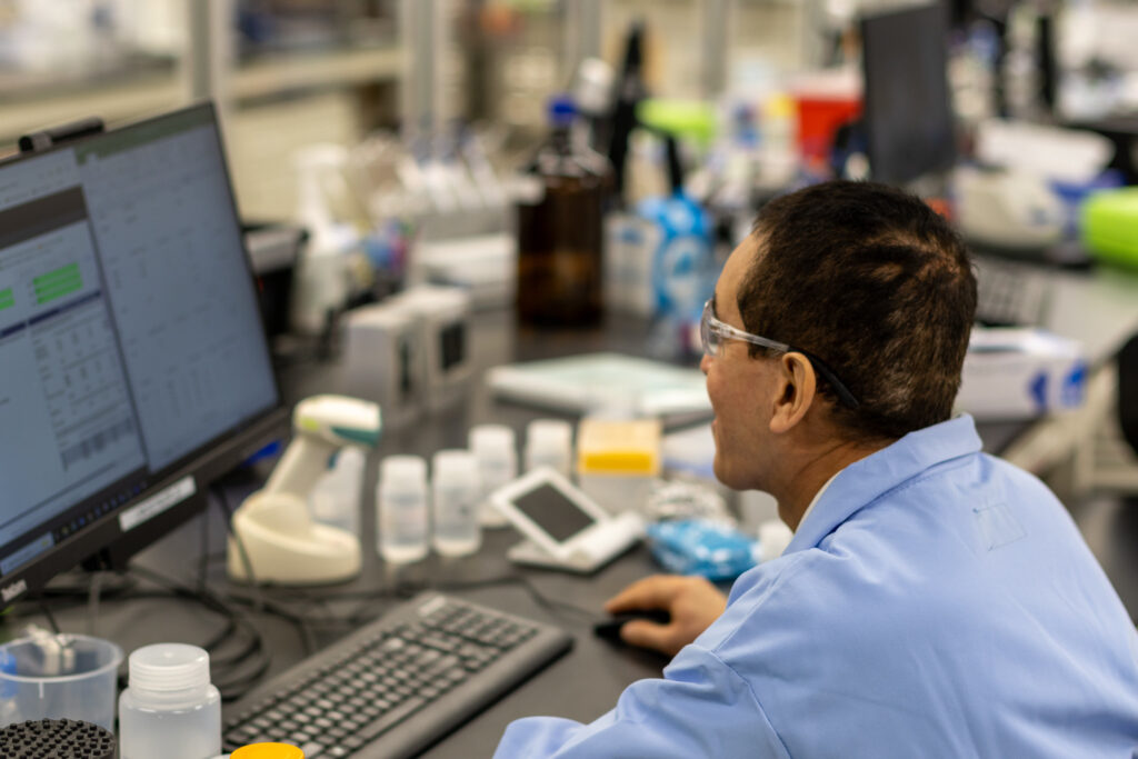 A man in a blue lab coat and safety glasses looks at a computer screen. His desk has lab equipment on it.