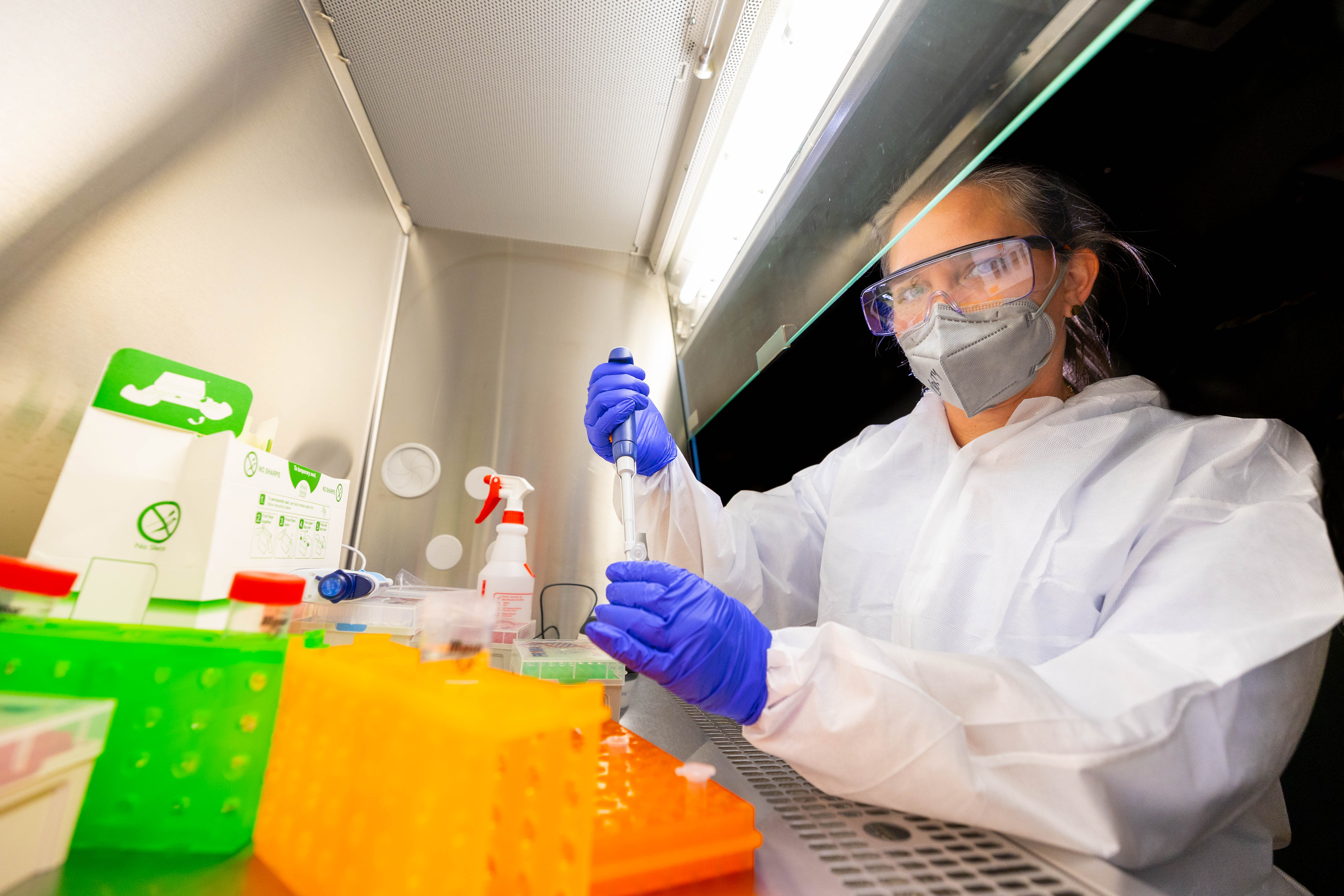 A woman in a lab coat uses a pipette to transfer liquid to a vial. She is surrounded by lab equipment.