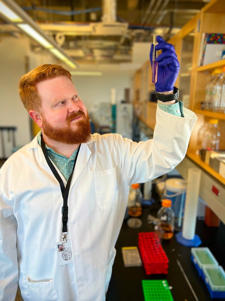 A man in a lab coat holds up a vial with purple liquid.
