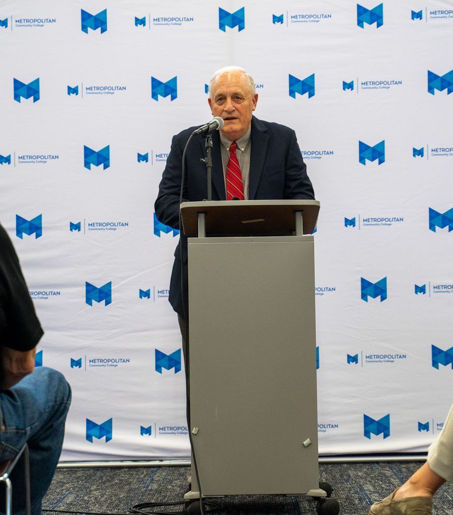 President Randy Schmaizl stands at a podium with a microphone. An MCC photo backdrop is behind him.